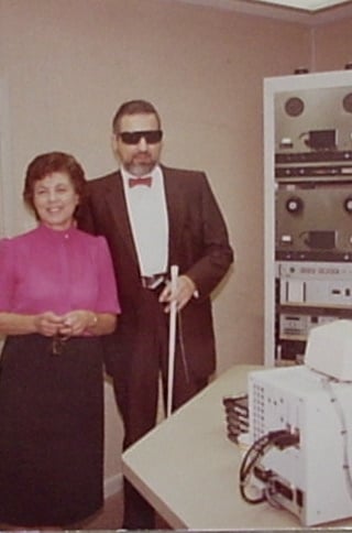 Dr. Robert McClean, the founder of WRBH, stands on the right wearing sunglasses and holding a white cane, dressed in a formal suit and bow tie. He is standing beside a woman in a pink blouse and dark skirt. Behind them is radio broadcasting equipment, including large machines and audio consoles.