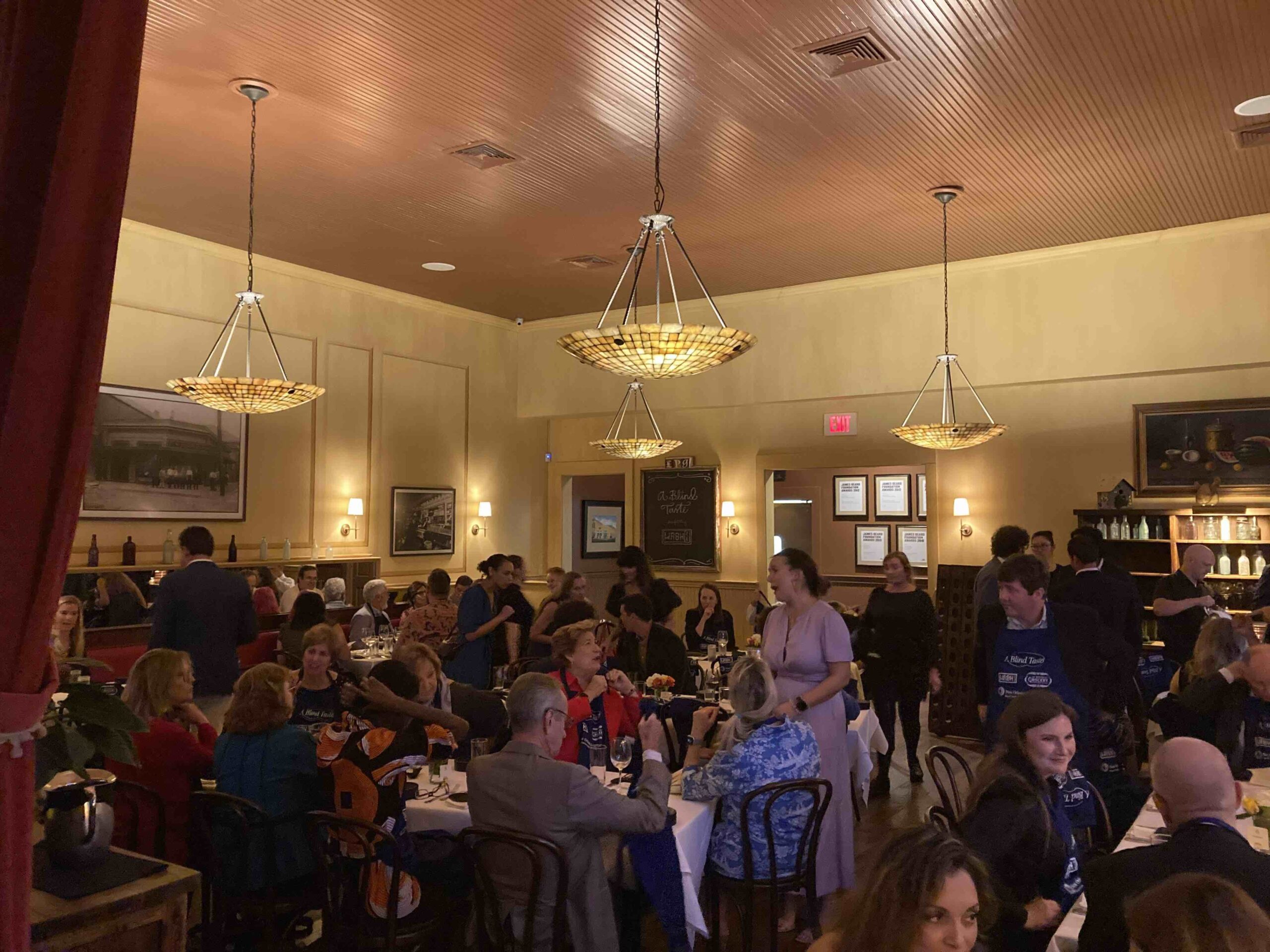 A wide shot of the dining room at A Blind Taste event, filled with guests wearing blindfolds and engaging in conversation. The room features elegant lighting with chandeliers, and the walls are adorned with framed artwork, creating a cozy ambiance.
