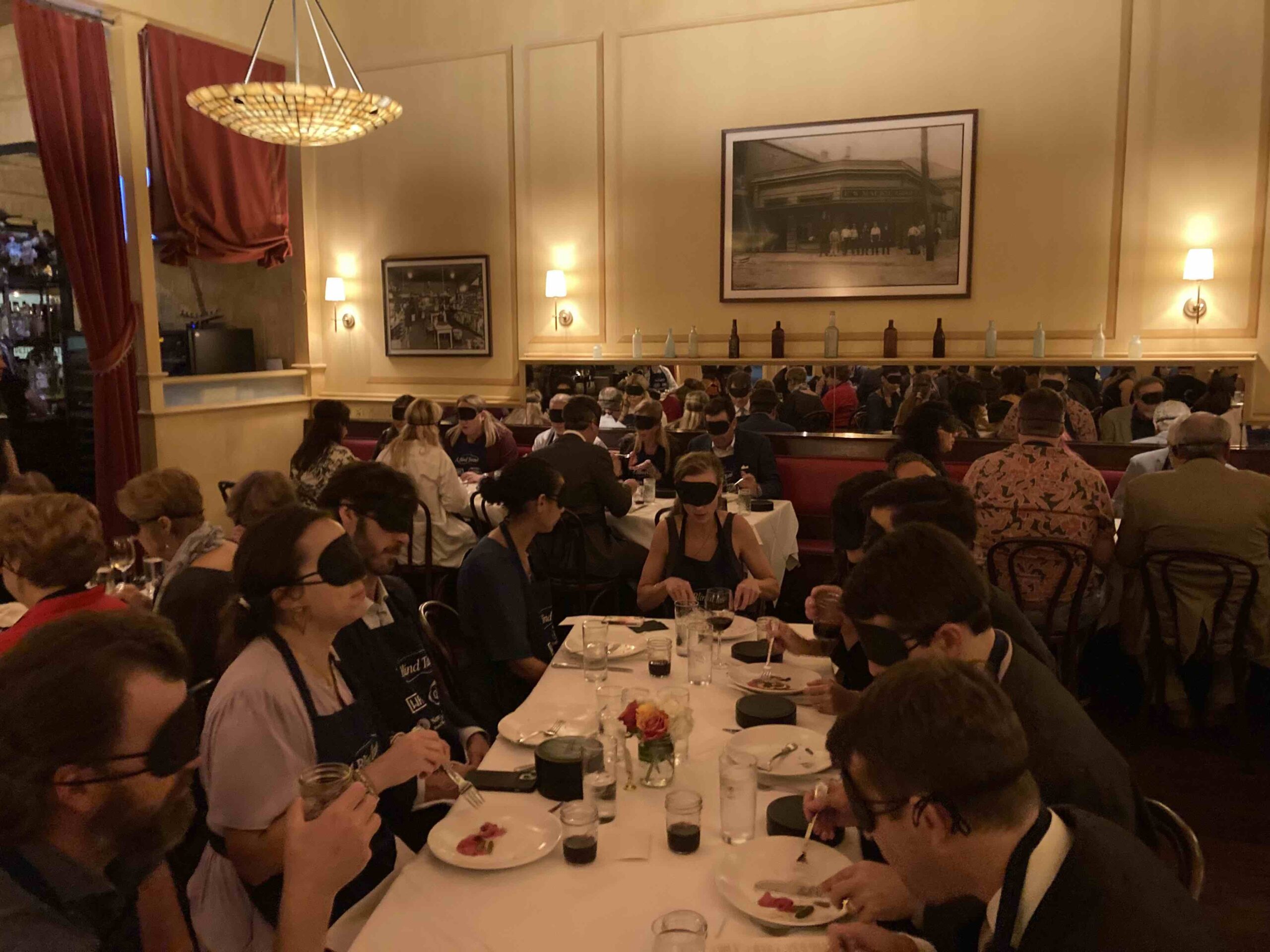 A view of A Blind Taste dinner, showing more attendees seated at tables, all wearing blindfolds while enjoying their meals. The intimate setting encourages conversation as guests explore the flavors of the dishes with heightened senses.