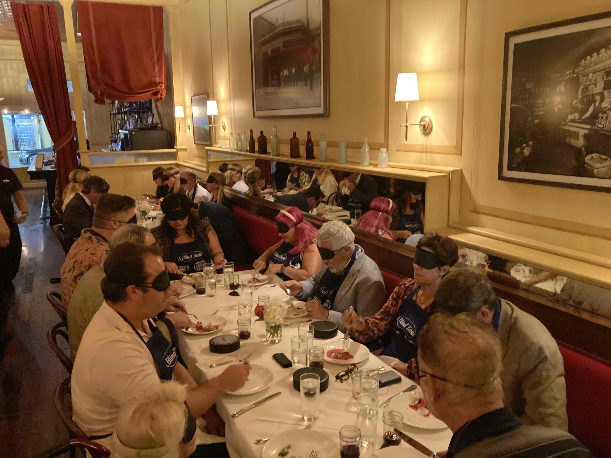 A close-up of several tables at A Blind Taste, with guests, wearing blindfolds, chatting and dining. The tables are filled with plates of food and drinks as everyone partakes in the unique sensory experience of dining without sight.