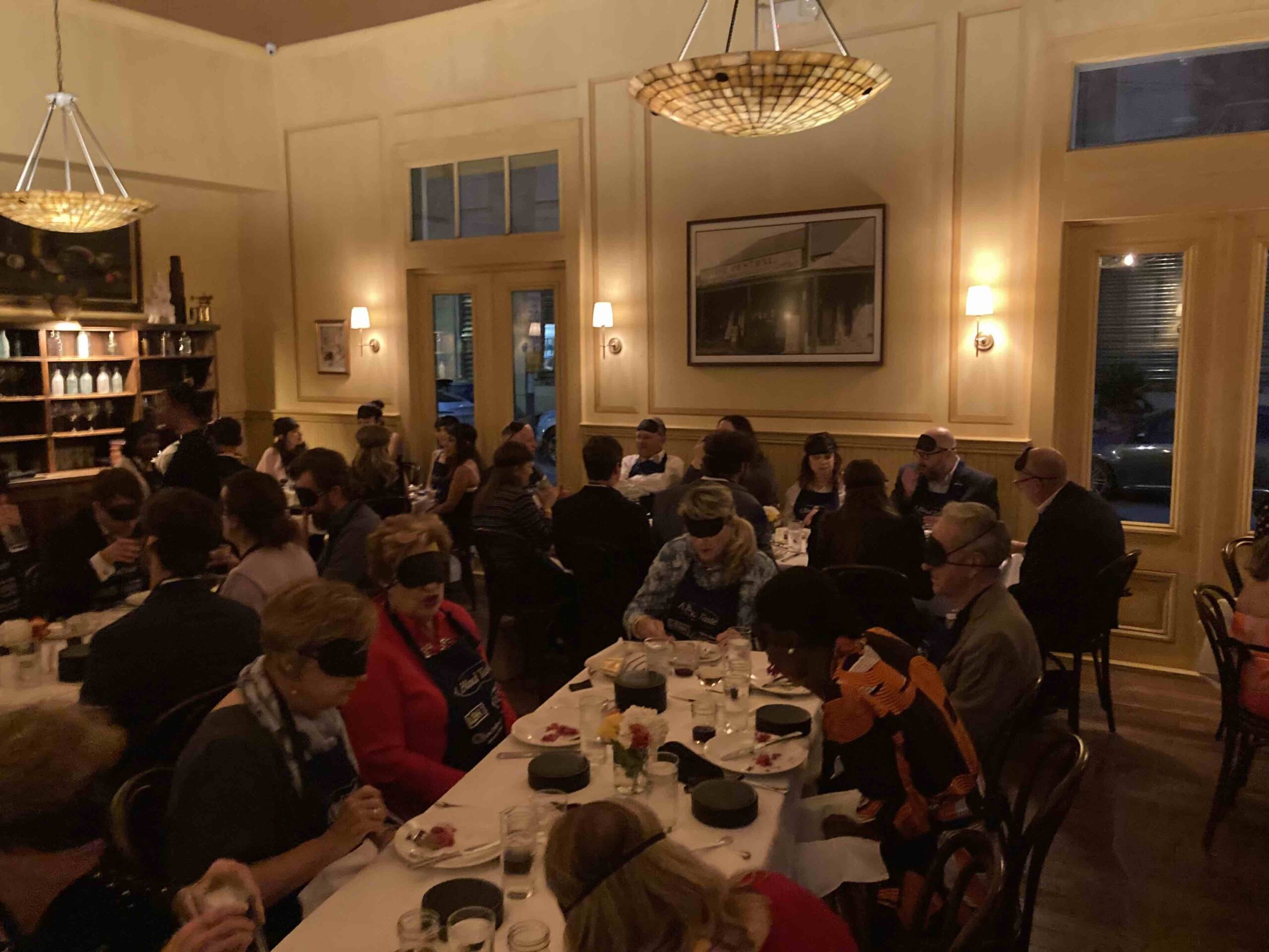 Attendees of A Blind Taste seated at long tables, dining while wearing blindfolds. The room is softly lit with warm lighting, and the atmosphere is intimate and engaging as guests enjoy their meals in a shared sensory experience.