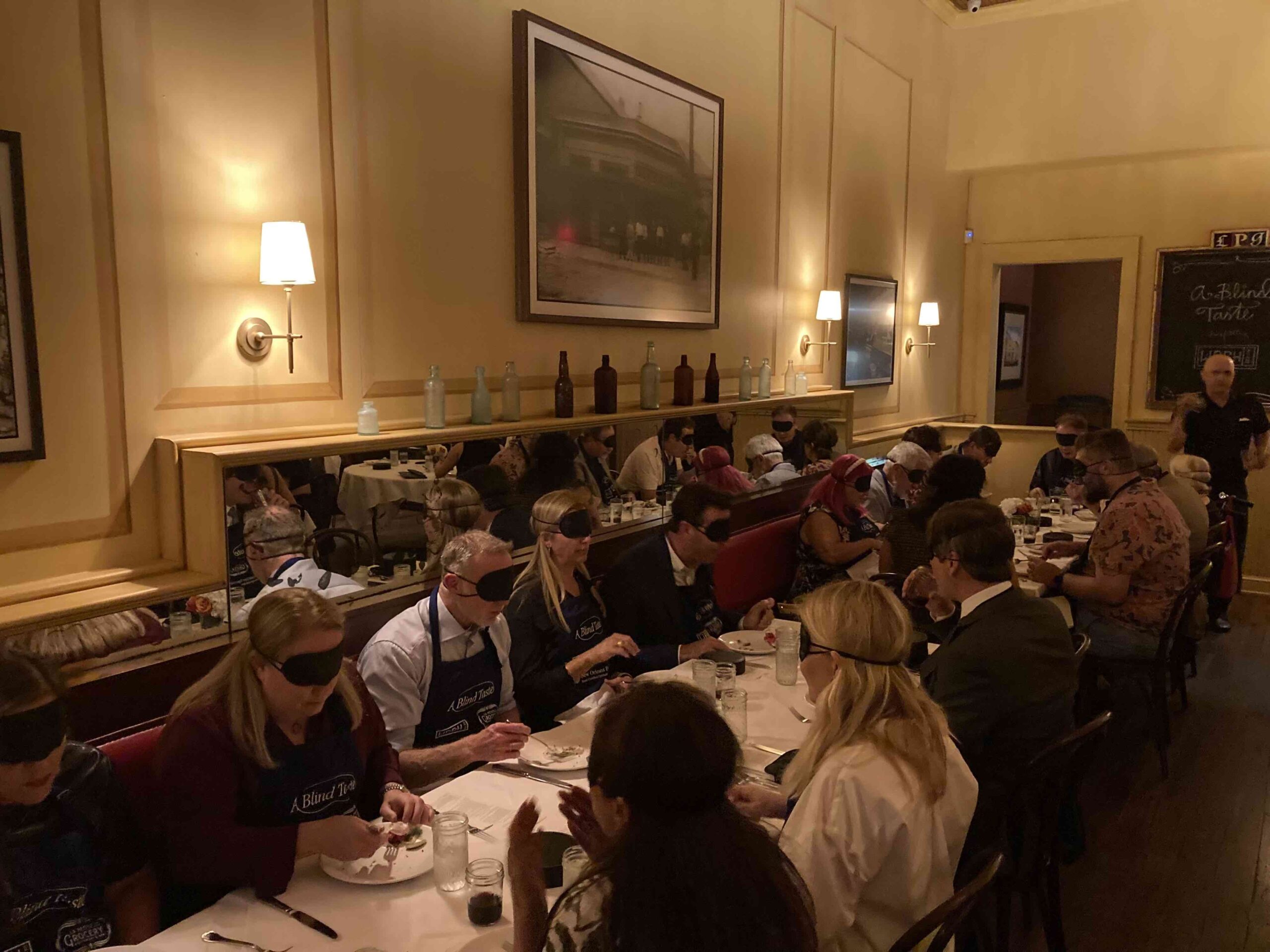 A wide shot of the event, showing rows of tables filled with guests participating in A Blind Taste. The diners, all blindfolded, are savoring their food in the elegant, warmly lit room, creating a reflective and engaging atmosphere.