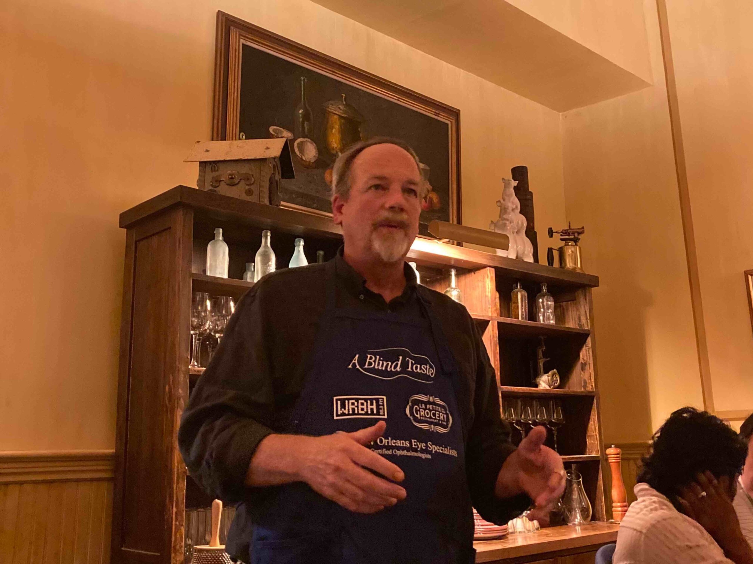 WRBH volunteer and wine presenter Geoff Worden standing in front of shelves of glassware at A Blind Taste event, wearing a dark shirt and apron with the event logo. He is addressing the attendees, with his hands raised as he speaks. Behind him is a painting and wooden cabinet.
