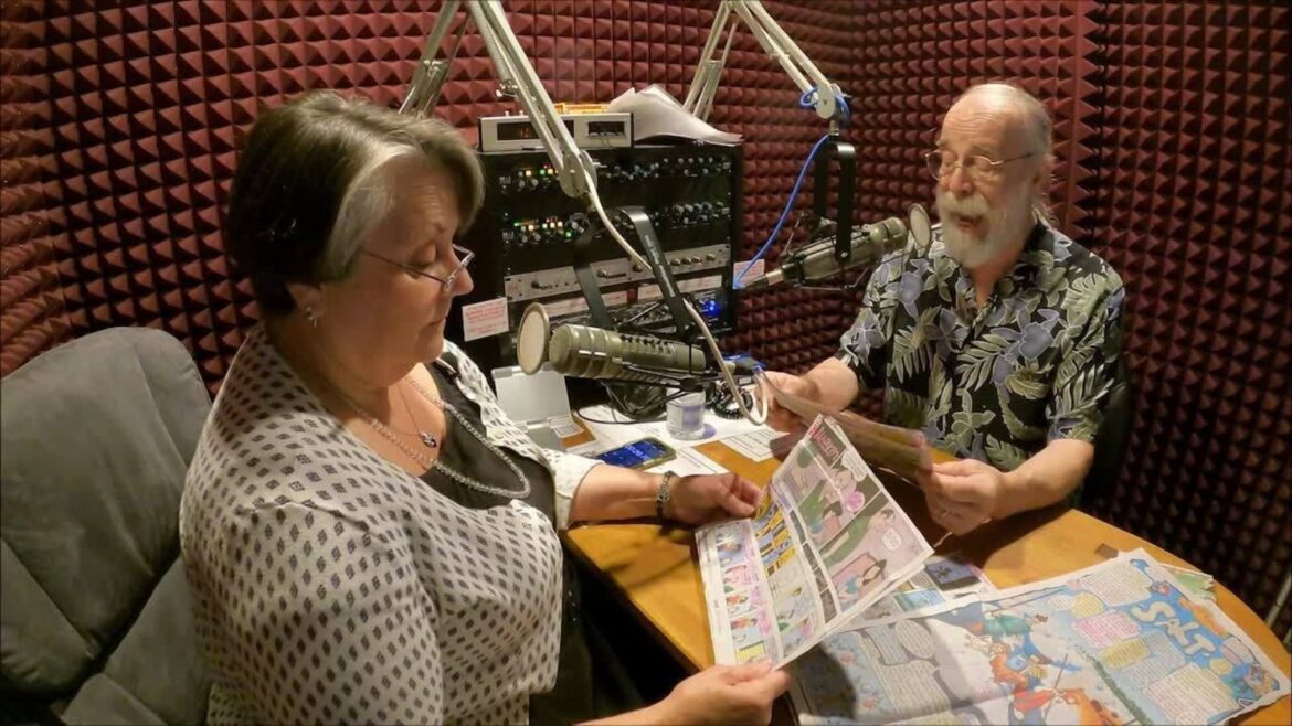 A man and a woman sit in a radio studio reading aloud from comic strips in front of professional microphones. The studio walls are lined with soundproof padding, and the table is covered with comic pages as they bring the stories to life for WRBH Reading Radio listeners.