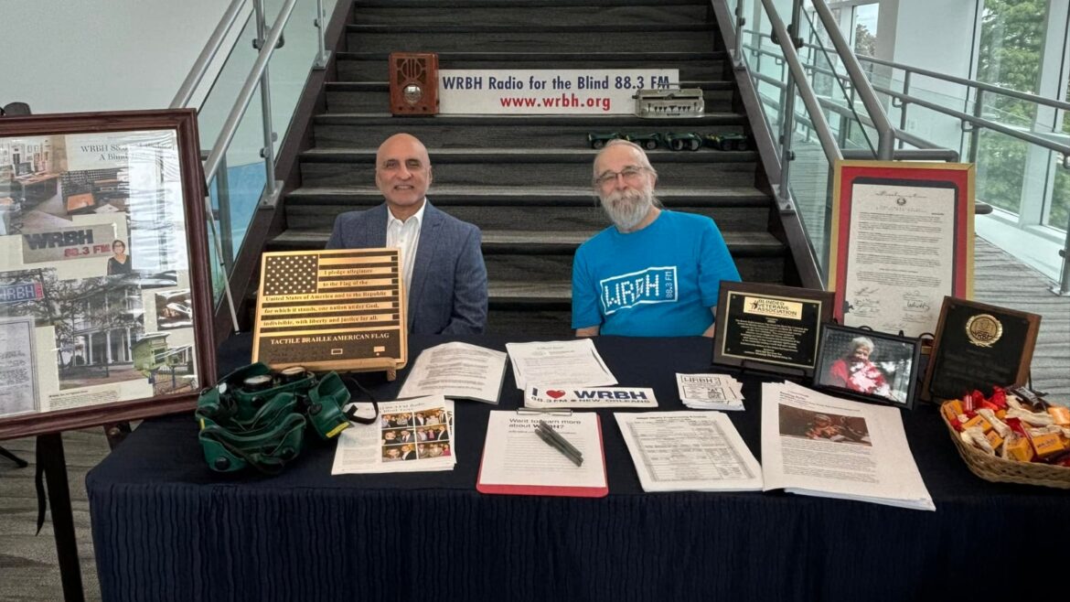 Two men, Carl Arredondo and Bruce Mohat, are seated at a booth for WRBH Reading Radio. Carl is wearing a gray suit and white shirt, while Bruce is wearing a bright blue WRBH T-shirt. They greeted attendees and spread the word about WRBH’s programming and services. The booth is set up in front of a large staircase with a WRBH banner hanging behind them that reads “WRBH Radio for the Blind 88.3 FM www.wrbh.org.” On the table are various items, including a Tactile Braille American flag, framed plaques, brochures, and documents promoting WRBH's services. A basket of snacks sits on the right side of the table.